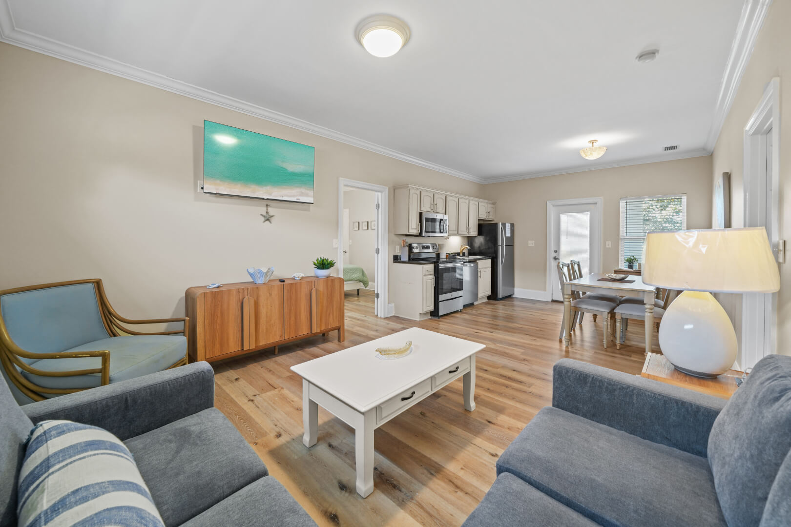 A bright and modern open-concept kitchen and living room with hardwood floors, white cabinetry, stainless steel appliances, a dining table with bar stools, and a cozy seating area with a sofa and TV. A large seashell artwork adds a decorative touch.